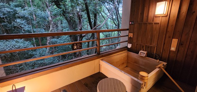A traditional Japanese wooden bath (ofuro) sits in a room with wood-paneled walls, next to a large window overlooking a lush green forest. A wooden ladle rests in the tub, and warm light emanates from a wall sconce.