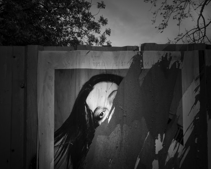 A moody black-and-white photograph of a striking mural depicting a haunting figure with a large eye, painted on a weathered wall beneath tree branches.