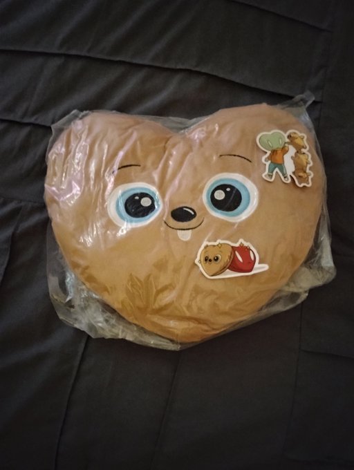 A heart-shaped brown plush pillow featuring a baby face design (with big blue eyes) and small bear stickers, packaged in clear plastic and resting on a dark gray bedsheet.