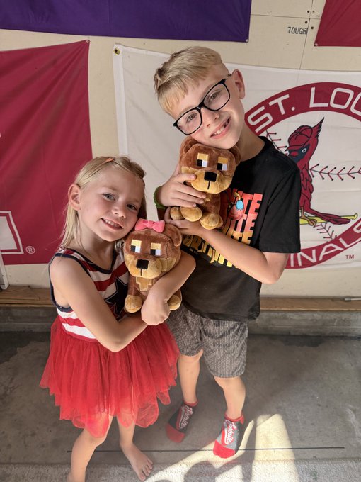 Two smiling children, a girl in a red tutu and a boy in glasses and a black shirt, pose together while holding Minecraft-themed dog plushies in front of a St. Louis Cardinals banner.