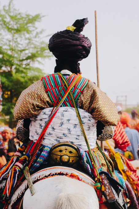A photograph showing the back of a person in vibrant, colorful traditional clothing and a dark turban, riding a horse.