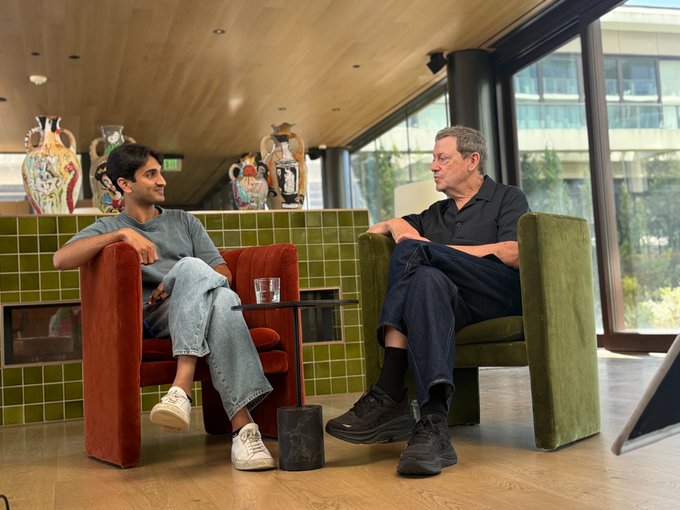 Two men sit in colorful armchairs having a conversation in a modern lounge with decorative ceramic vases, large windows, and a green-tiled wall, captured in a relaxed interview-style setting that suggests an upcoming podcast or discussion episode tied to the "coming soon" message.