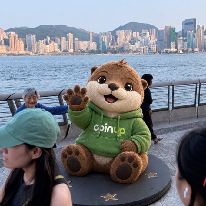 A large otter mascot statue wearing a green 'coinup' hoodie sits on a waterfront promenade waving, with Hong Kong's skyline and harbor in the background.