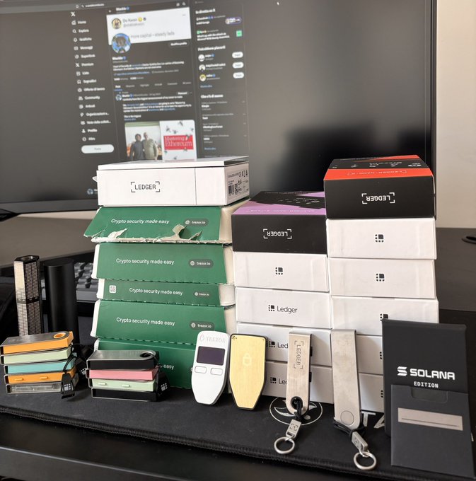 A collection of cryptocurrency hardware wallets, including Ledger devices and Trezor, arranged in front of boxes with their original packaging (including a "Solana" edition) on a desk with a monitor in the background.