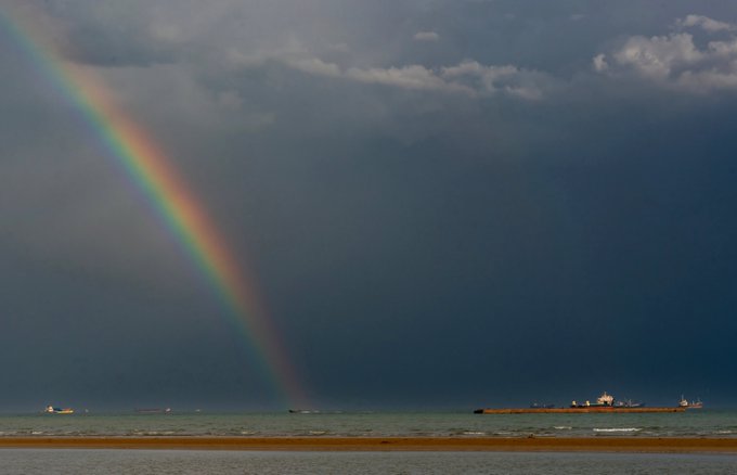 A vibrant rainbow arcs dramatically across a stormy, cloud-filled sky over a calm sea, with small boats visible on the horizon near a distant shoreline structure. The scene beautifully captures the contrast between dark, moody weather and the colorful optical phenomenon, complementing the post's theme of atmospheric ocean photography.