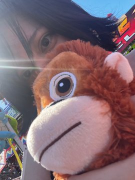 A person smiles for a selfie while holding a plush monkey toy with a fuzzy orange body and a smiling face. In the background, a colorful carnival ride can be seen under a bright blue sky.