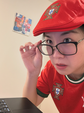 A woman wearing a red Portugal soccer jersey and matching cap adjusts her glasses while posing in front of a laptop, ready to cheer on Portugal in their match against Croatia on the OKX World Cup outcome market.