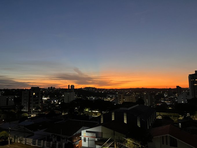 A cityscape at dusk captures a vibrant orange and blue sunset over a sprawling urban area, with buildings and distant hills silhouetted against the colorful sky.