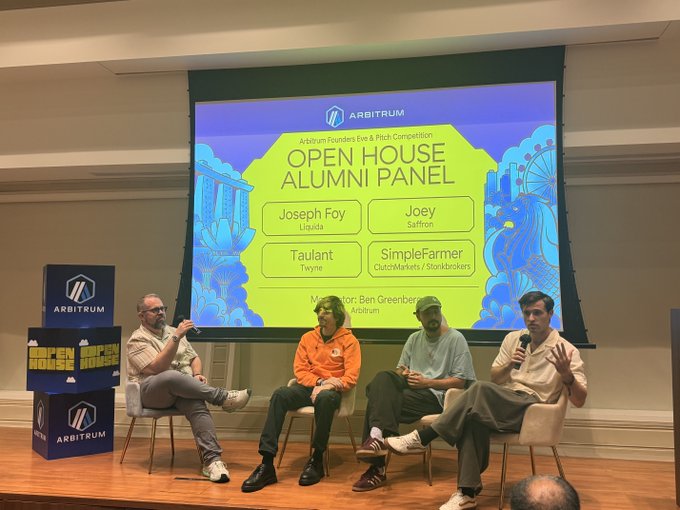 An 'Open House Alumni Panel' event hosted by Arbitrum, featuring four speakers (Joseph Foy, Joey, Taulant, SimpleFarmer) seated on stage with moderator Ben Greenberg.