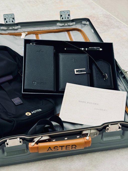 An open ASTER-branded suitcase containing a black leather wallet set (wallet, cardholder, and luggage tag), a black ASTER bag, and a white card with Chinese text reading '每段旅程，都是生活限定。Aster陪你路上。'.