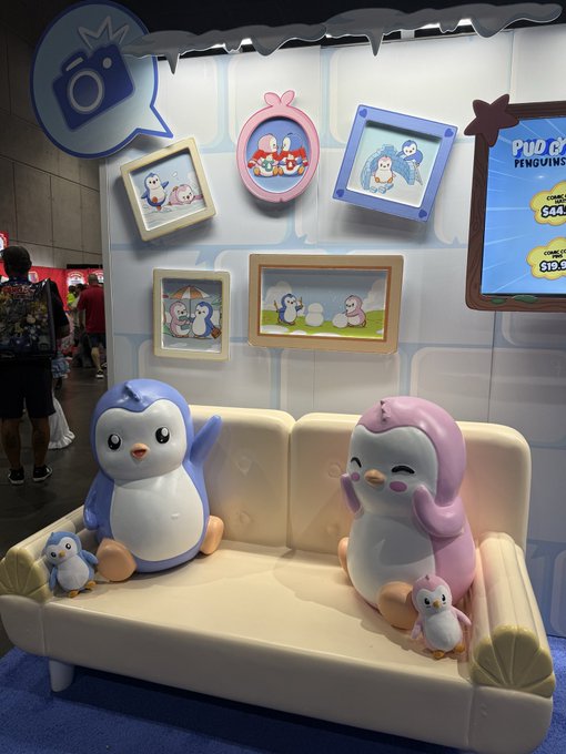 A convention booth display featuring cute penguin figurines and framed pictures of 'Pudgy Penguins,' with a screen advertising 'PUDGY PENGUINS' plush toys starting at $19.99.