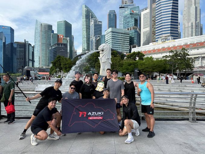A group of men poses for a photo in front of the Merlion statue in Singapore, holding a black banner that reads 'AZUKI WEB3 CLICKS SG EDITION' with a red logo.