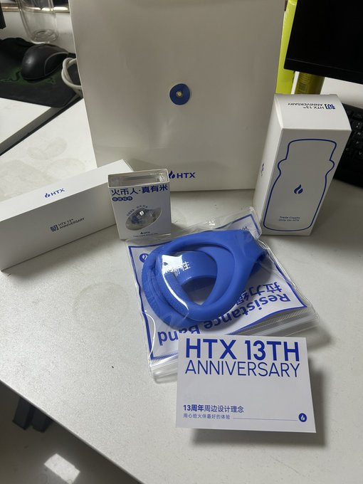 A collection of HTX 13th Anniversary promotional merchandise is displayed on a desk, including a white paper bag, branded boxes, and a blue resistance band accessory.