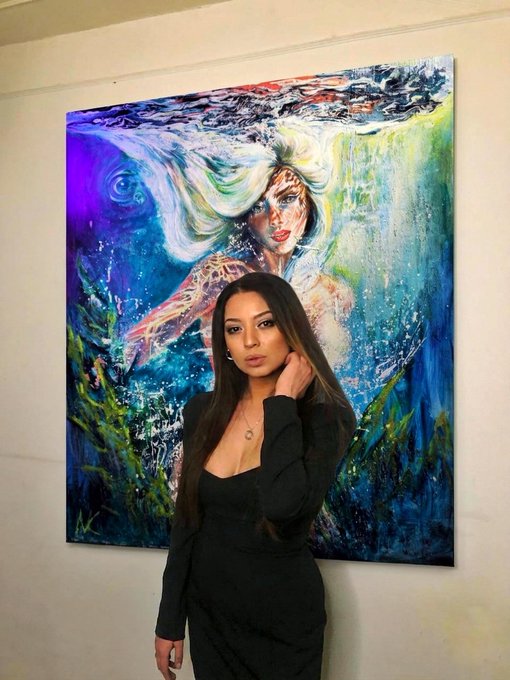 A woman in a black dress poses in front of a large, vibrant abstract painting depicting an underwater mermaid scene with flowing hair.