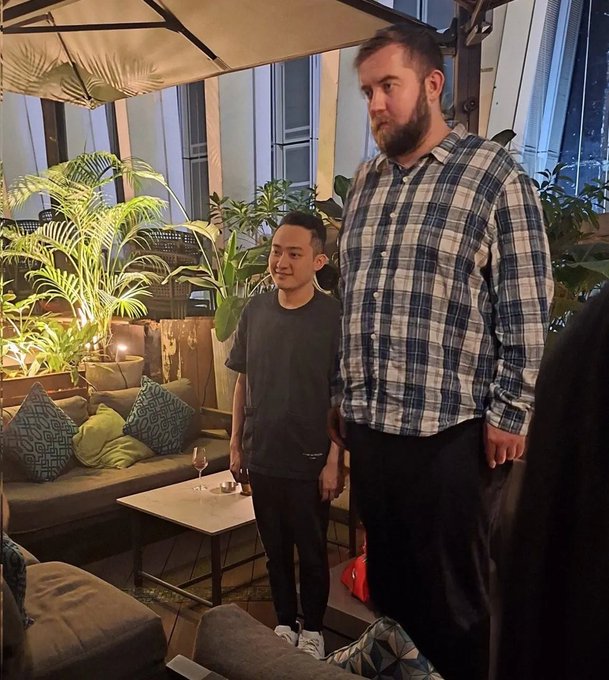 Two men pose for a photo at an outdoor lounge area surrounded by tropical plants, with one wearing a black t-shirt and the other in a blue and white plaid shirt. The casual, social setting suggests a gathering or event, which aligns with the promotional post announcing a new episode of "Linn's Leverage" featuring crypto-related platforms.