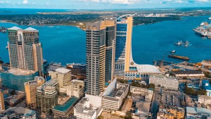 An aerial cityscape view showing tall skyscrapers, a waterfront with ships and cranes, and a distant shoreline under a partly cloudy sky.