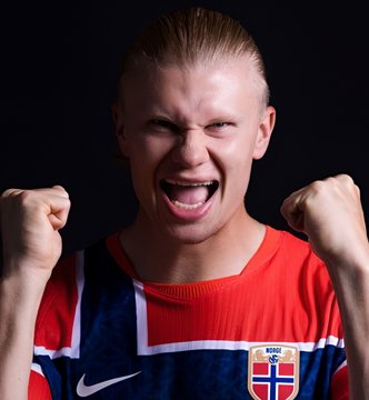 A young man with blonde hair, mouth wide open in an intense celebratory shout and fists clenched in triumph, wears a red Norway national football team jersey featuring the NB crest and Nike logo, capturing the passionate moment tied to the post's discussion of Norway's World Cup 2026 prospects ahead of their match against France.