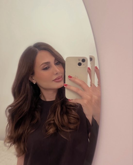 Mirror selfie of a woman with long wavy brunette hair wearing a black top, holding a white iPhone with triple camera array, featuring red manicured nails and subtle makeup, photographed against a pink and white wall.