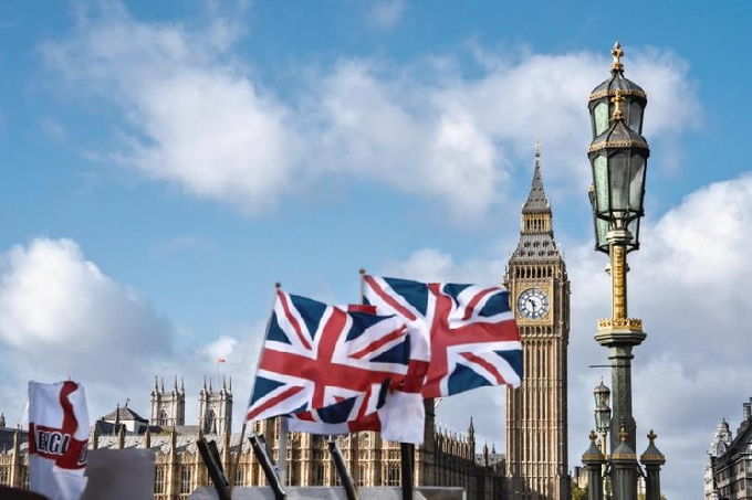 Two Union Jack flags wave in front of Big Ben and the Houses of Parliament in London, with an ornate street lamp in the foreground against a partly cloudy sky.
