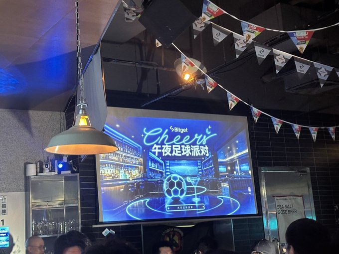 A large screen displays "Bitget Cheers 午夜足球派对" (Football Night Party) with neon designs of a soccer ball and beer glass, surrounded by colorful Bitget pennant banners hanging from the ceiling of a lively bar, matching the post's invitation-only atmosphere to Bitget's sponsored football viewing party.