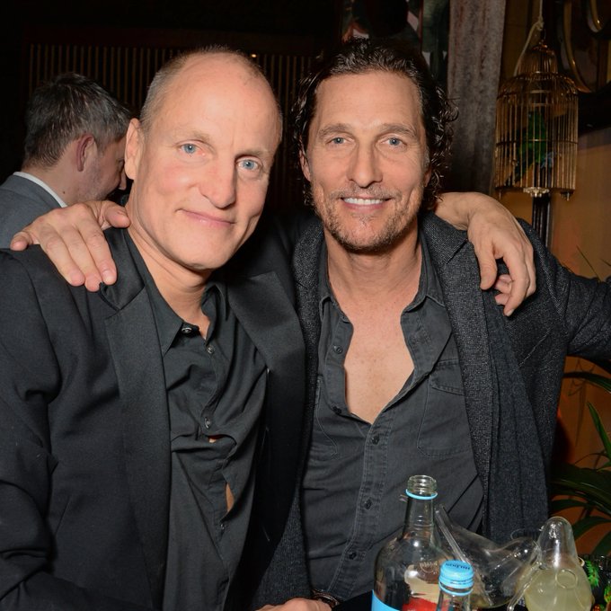 Two men pose with arms around each other at what appears to be a social event, with bottles and a glass visible in the foreground. The image relates to speculation about whether the two actors—Woody Harrelson and Matthew McConaughey—might be half-brothers, as Harrelson recently claimed his father had a relationship with McConaughey's mother.