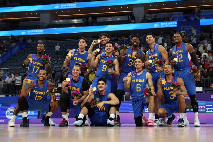 The Philippine men's basketball team poses together on a podium, proudly wearing blue 'PILIPINAS' jerseys and displaying their gold medals, likely after winning the 2023 Asian Games.
