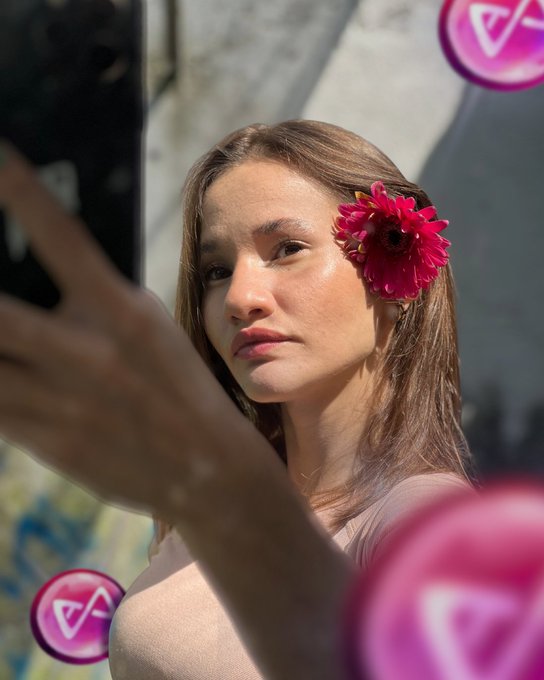A woman with a vibrant magenta flower in her hair, with out-of-focus purple Valentine's-themed emoji bubbles in the foreground.