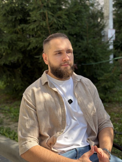 A portrait photo of a bearded man with short hair wearing a beige corduroy shirt over a white t-shirt, sitting outdoors with green trees in the background.