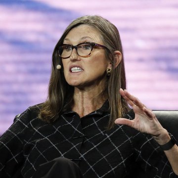 Cathie Wood, CEO of ARK Invest, is shown speaking on stage with an animated expression and hand gesture. The image accompanies news about her firm's launch of a tokenized venture fund on the Ethereum blockchain, allowing 24/7 trading exposure to ARK's portfolio of private and public companies.