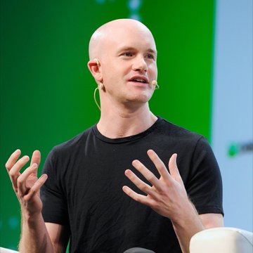A bald man in a black t-shirt gestures while speaking into a headset microphone against a green backdrop. The image relates to Coinbase's launch of new AI tools for agentic trading, commerce, and payments as the company expands infrastructure for the emerging agentic economy.