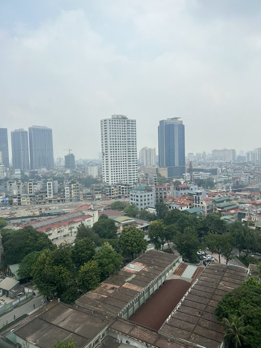An elevated view of a hazy city skyline with modern high-rise buildings towering above lower residential structures and a tree-lined foreground.