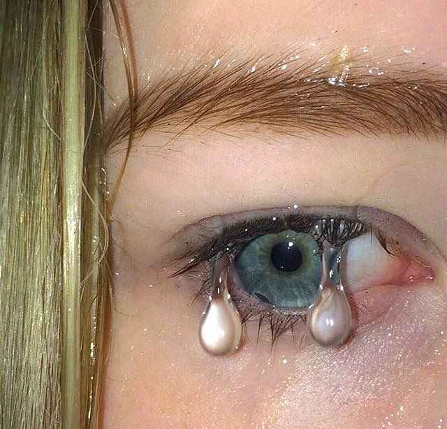 A hyper-realistic digital artwork showing an extreme close-up of a person's eye with teal irises, shimmering glitter-decorated eyebrows, and two large pearls hanging like tears from the eyelashes.
