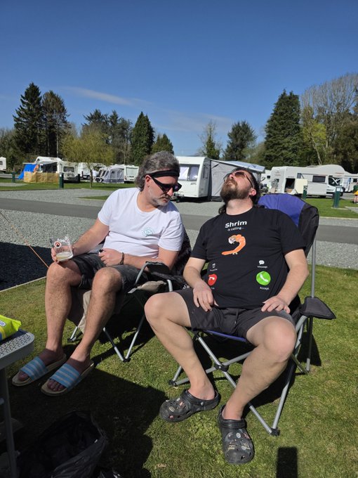 Two men relaxing in camping chairs outdoors; the left one has long grey hair and sunglasses, while the right wears a black "Shrim" t-shirt with croc shoes. Caravans and trees are visible in the background.