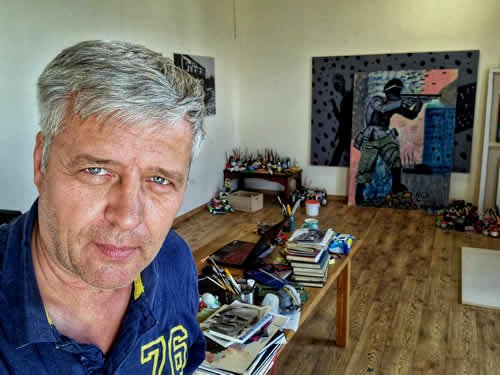A middle-aged man with gray hair takes a selfie in an art studio, with paintings, art supplies, and a wooden floor visible behind him.