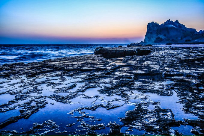 A coastal landscape at sunset featuring wet rocky tide pools in the foreground, ocean waves, and an iceberg visible on the horizon under a gradient sky.