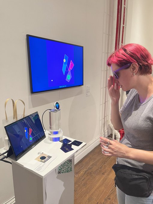 A person with pink hair wearing purple sunglasses examines an interactive display featuring a screen and a small device on a white pedestal.