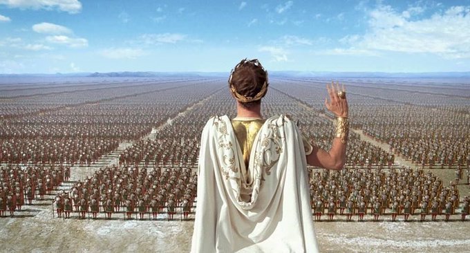 The image features a man standing in front of an expansive desert landscape, with a large number of people visible in the background. The man appears to be wearing a white robe or cape, which stands out against the desert backdrop. He is waving his hand towards the camera and looking off into the distance. The scene suggests that this could be a historical event or a significant moment captured in time.