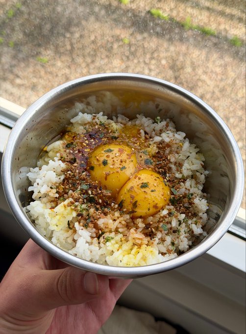 A hand holds a metal bowl filled with white rice topped with three raw egg yolks, red chili flakes, seaweed, and seasoning.