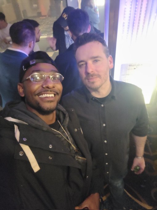 Two men pose for a photo at what appears to be a social event; the man on the left wears a black cap, glasses, and jacket, while the man on the right wears a dark button-up shirt.
