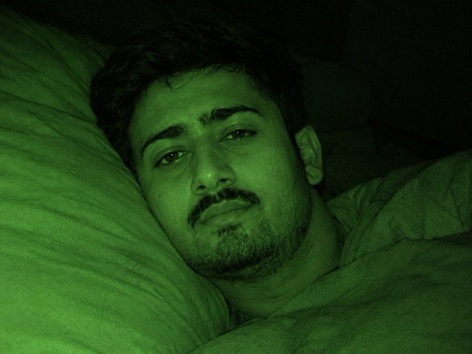 A man with a beard lying in bed in a dark room, captured with a strong green night-vision tint.