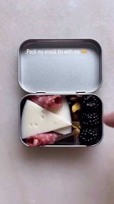 A small metal tin is packed with cheese triangles, salami slices, almonds, and fresh blackberries for an on-the-go "milady dinner," as captioned by the overlay text "Pack my snack tin with me 🧀."