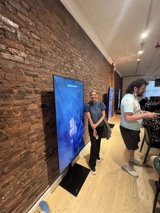 Photo of a woman with short gray hair standing next to a digital display screen showing an ocean image, with 'Stellar Vacation' text, in a room with exposed brick walls.