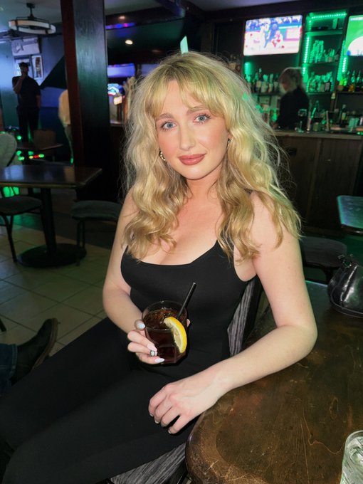 A young woman with long blonde hair and a black top sits at a bar, holding a glass with a dark beverage and a lemon slice.