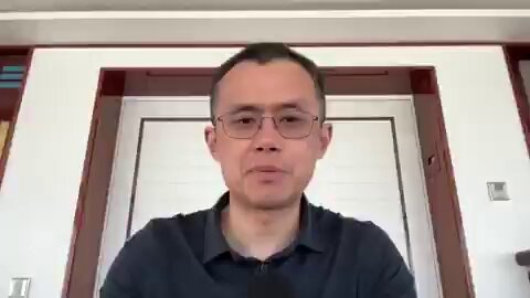 The image shows a man wearing glasses sitting at a desk with his hands on his lap. He appears to be looking directly into the camera while talking. The background of the image features a white door, suggesting that he might be in an office or workspace setting.