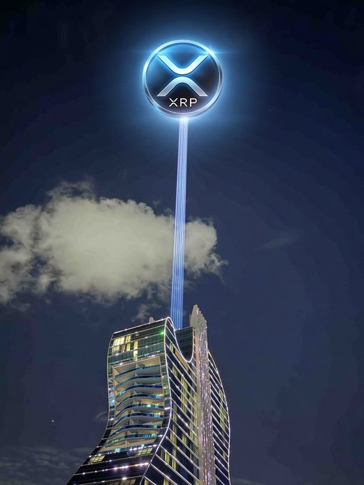 A glowing blue XRP cryptocurrency logo hovers above a skyscraper at night, connected to the building by a beam of light passing through a cloud. The image visually represents the post's anticipation of an upcoming XRP and XLM event, with the illuminated logo symbolizing a high-profile "show" being staged in the crypto world.