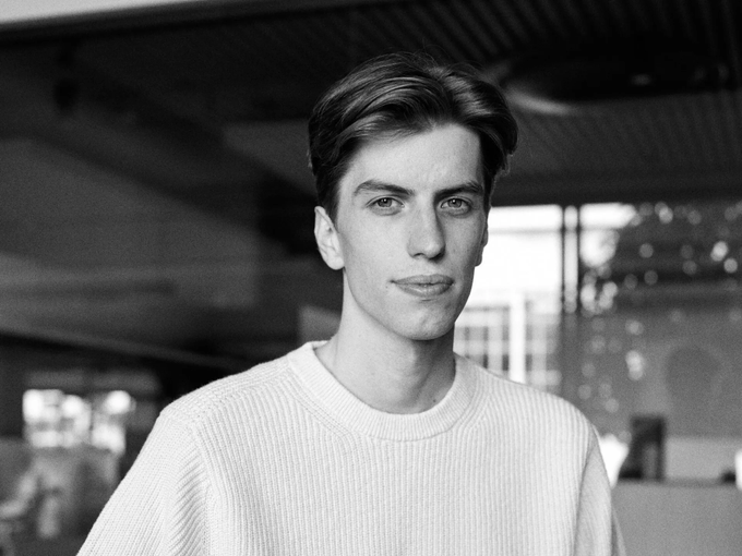 A young man with neatly styled hair and a white sweater gazes confidently at the camera in a black-and-white portrait. The image likely represents the speaker in their youth, evoking a sense of fresh ambition and the early stages of a career in finance and trading.