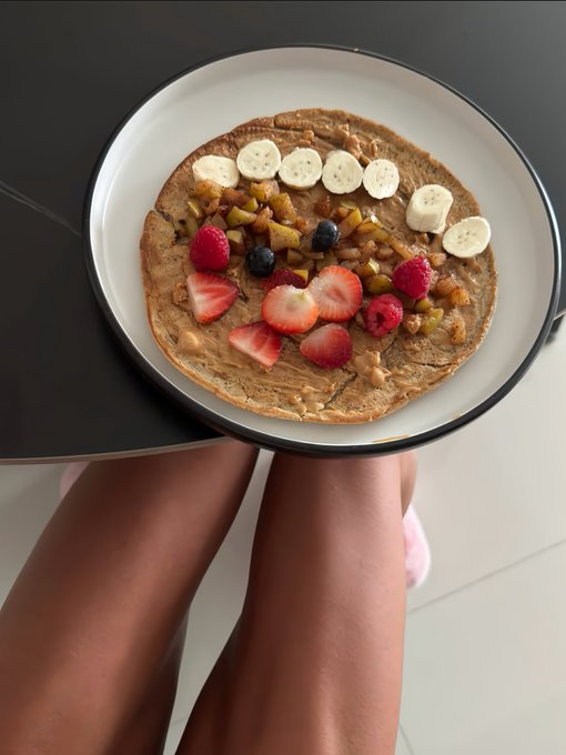 A person holds a plate containing a pancake or crepe topped with peanut butter, banana slices, strawberries, raspberries, and blueberries.