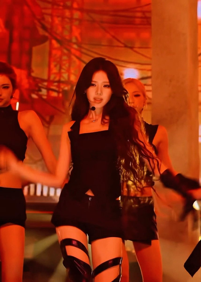 A female performer with long dark hair dances on stage in a black outfit with thigh straps, flanked by backup dancers against a red backdrop.