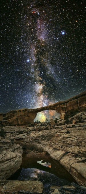 A star-filled night sky, dominated by the Milky Way, glows above a rugged desert canyon and reflective pool. The dramatic cosmic scene complements the post’s theme, “Cosmos.”