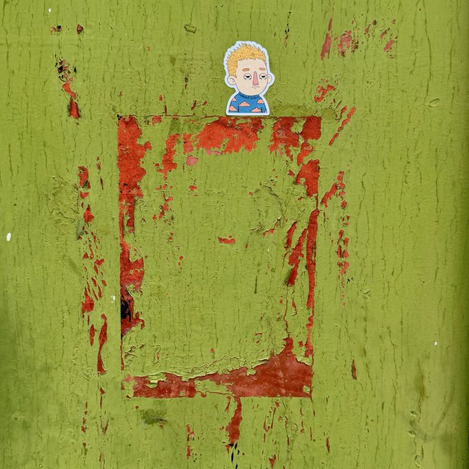A cartoon sticker of a blond-haired person wearing a blue cloud-patterned shirt is placed on a peeling green surface with exposed red underlayer. The image shows the result of printing stickers, fulfilling the post's hope.